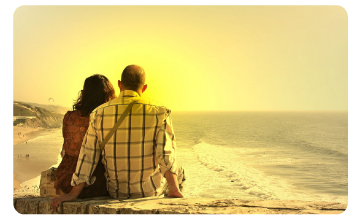 A couple sitting close together on a cliff edge, watching the sun set over the ocean.